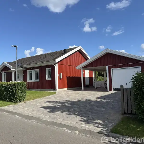 Et rødt hus med hvide vinduer står ved en brolagt indkørsel. Ved siden er en carport med hæk omkring grunden. Himlen er blå med få skyer.