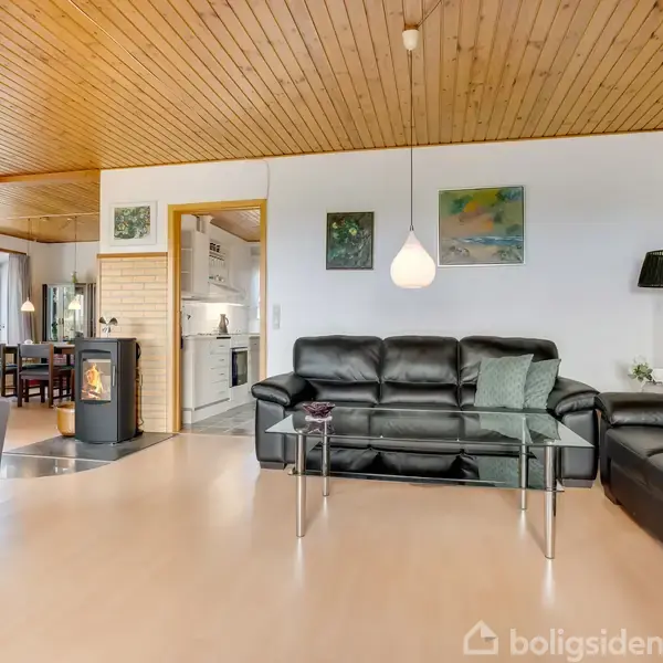 Sort lædersofa står stille i lys stue med trægulv og loft, omgivet af glasbord, malerier på væggen og stor lampe, nær en spisestue med brændeovn.