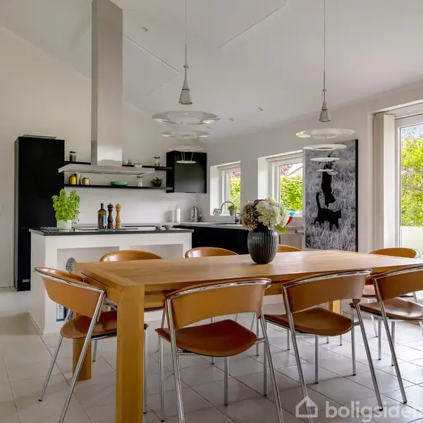Et spisebord står i midten omgivet af seks stole; i baggrunden ses et moderne køkken med kogeplade. Store vinduer og en glasdør giver udsigt til en grøn have.