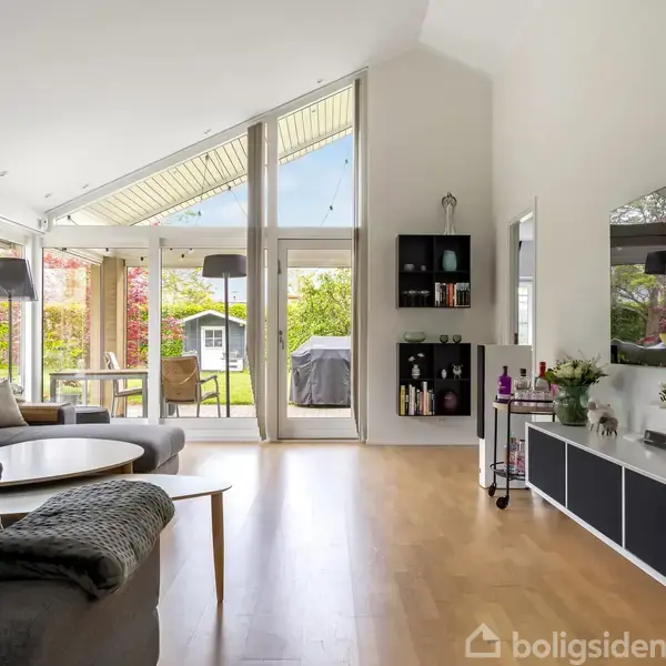 Stue med grå sofa, lave sofaborde og stor fladskærm på væggen. Store vinduer åbner mod en terrasse med udsigt til en grøn have. Reol med dekorative genstande ved siden af fjernsynet.