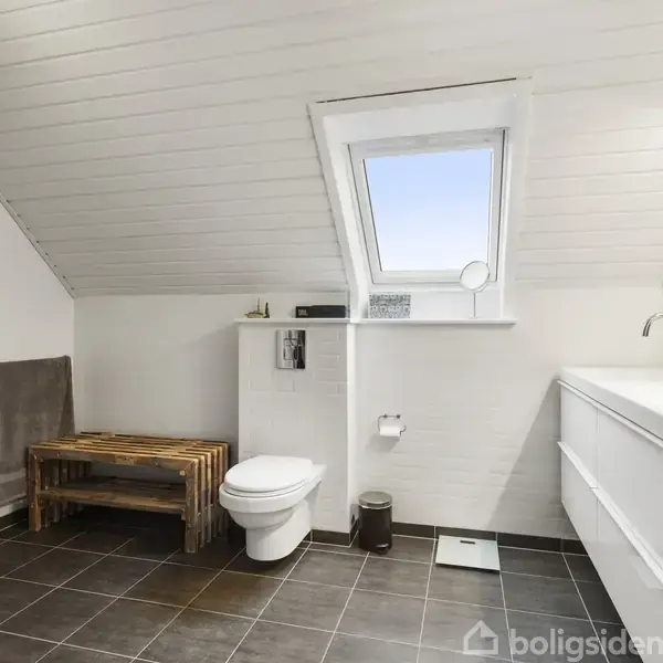 Badeværelse med toilet placeret under et skråt loft. Træbænk og håndklæde til venstre, vask med spejl til højre. Lyse fliser og et vindue over toilettet.