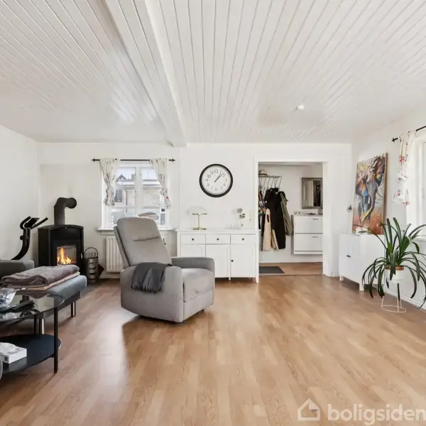 Stue med grå sofa, lænestol, brændeovn, vægur og træbord. Indretningen er lys med trægulv og hvide vægge. Store vinduer giver naturligt lys, og en plante står ved siden af en kommode.