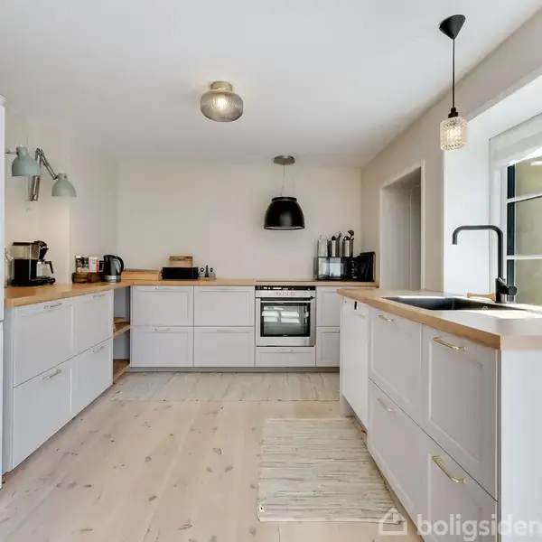 Køkken med hvide skabe, træbordplader, og hvidevarer, herunder køleskab og ovn. Vindue til højre giver naturligt lys. Lampe over vasken. Lys trægulv med lille tæppe.
