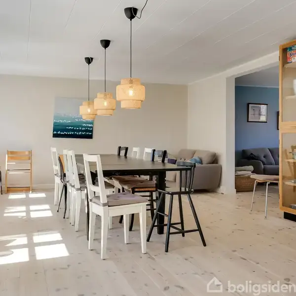 Spisestue med rektangulært spisebord og stole, belyst af fire hængelamper. Vinduer til venstre giver naturligt lys. Glasskab til højre, sofa i baggrunden gennem en døråbning.