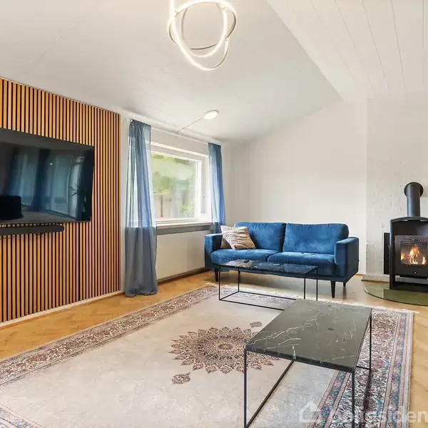 Fladskærms-TV hænger på lamelvæg; rummet har blå sofaer, brændeovn og tæppe; stue med træparketgulv og store vinduer med blå gardiner.