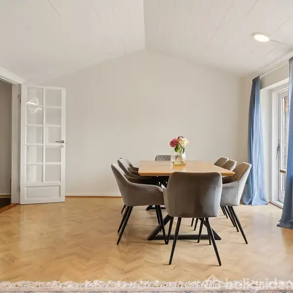 Spisebord med seks stole står i en lys stue. Blomster i en vase pryder bordet. Store vinduer med blå gardiner giver udsigt til omgivelserne. Tændte loftlys ses.