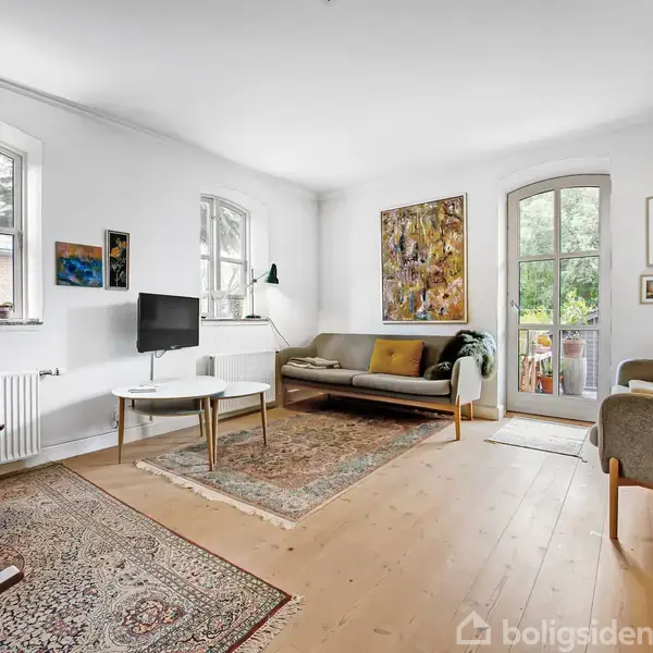 Stue med to sofaer placeret omkring et rundt sofabord, dekoreret med farverige tæpper og kunst på væggene. Lyse vægge og store vinduer giver et lyst og indbydende udtryk.