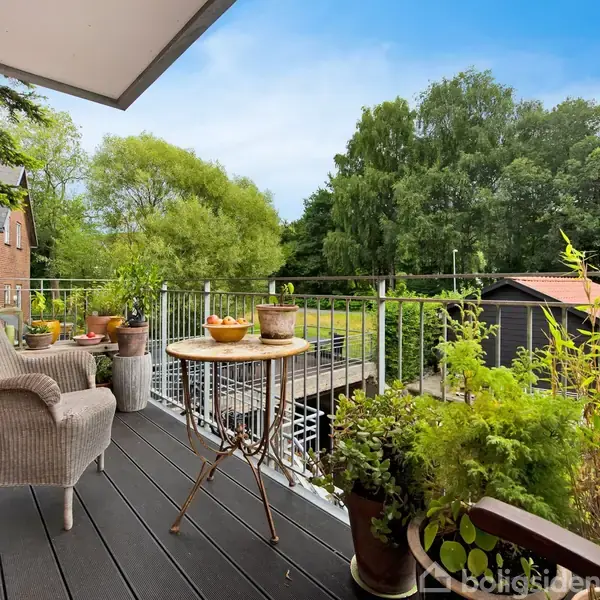 Terrassemøbler står stille; en flettet stol og et lille bord. Omgivet af planter og udsigt over en frodig have med træer og en lille hytte i baggrunden.