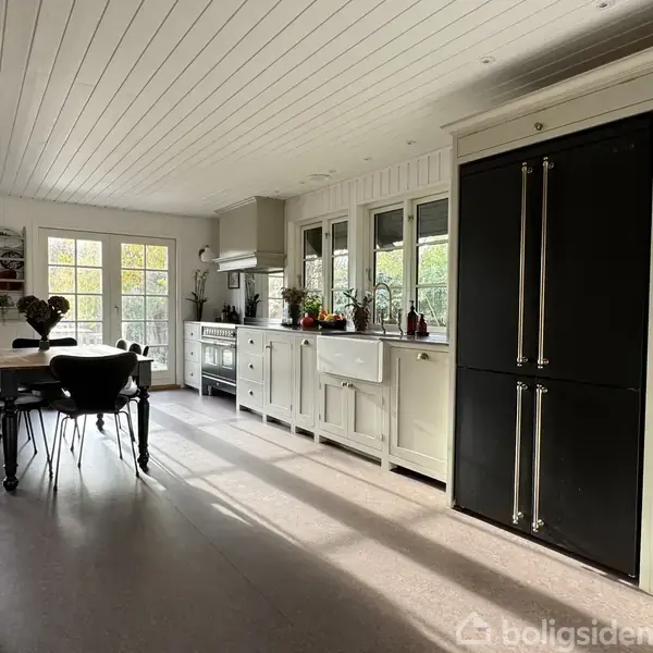 Et moderne køkken med et stort sort køleskab, hvide skabe og en vask ved vinduerne. Et spisebord står i nærheden, oplyst af sollys fra dobbeltdøre.