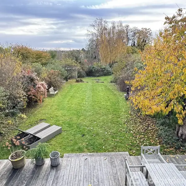 Grøn græsplæne strækker sig gennem en lang have; efterårsfarvede træer og buske omkranser den. Foran ses terrasse med havemøbler og krukker, under en overskyet himmel.