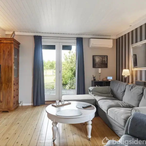 En stue med grå sofa står foran et rundt, hvidt sofabord. Et fladskærms-TV hænger på væggen. Rummet har trægulv, stribede vægge og en stor dør med udsigt til grønne omgivelser.