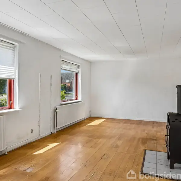 Brændeovn står stille i en lys stue med hvide vægge og trægulv. Vinduer med gardiner slipper sollys ind, og en radiator er placeret under vinduet. Ingen tekst fundet.