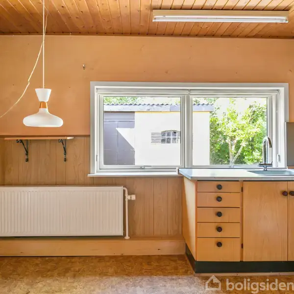 Et køkken med træloft og -skabe, har en lampe hængende over en radiator. Et vindue ved vasken giver udsigt til en gård med en bygning og træer.
