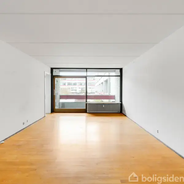 Stort, ryddeligt rum med trægulv, der åbner ud mod en altan gennem store glasdøre. Hvide vægge og et tilstødende rum kan ses gennem en åben døråbning til venstre.