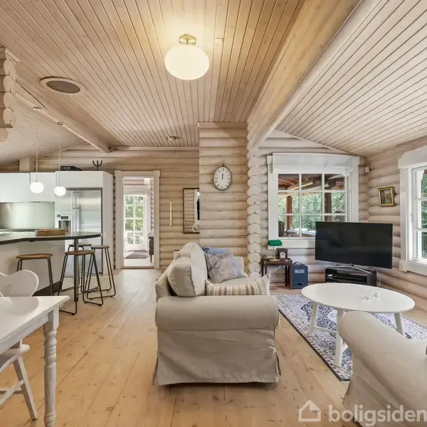 En lys stue med trævægge og loft, sofaer mod et stort vindue, et rundt sofabord og et TV. Til venstre ses et spisebord, og i baggrunden er et køkken med barstole.