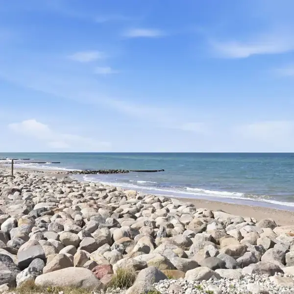 Store sten ligger på en strand og strækker sig langs kystlinjen, ledsaget af bølger der forsigtigt ruller ind mod land. I baggrunden ses en blå himmel med hvide skyer.