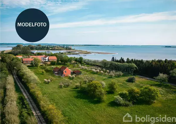 Modelfoto af et rødt hus omgivet af grønne marker og træer, med udsigt til hav og kystlinje i baggrunden under en klar himmel.