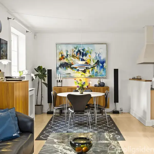 Et moderne opholdsrum med sort sofa, marmorbord og stol. Rundt spisebord med blomsterbukket i lys stue med kunst på væggen og træskab. Åben adgang til køkkenet.