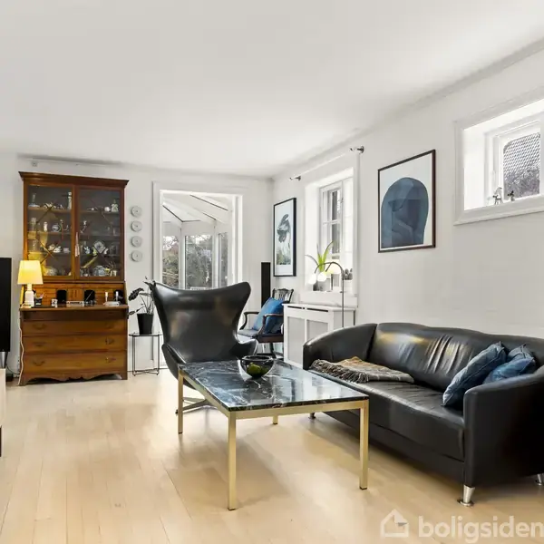 En sort lædersofa står i en lys stue med trægulv. Foran sofaen er et sofabord, og i baggrunden ses en reol med glaslåger og diverse dekorationer.