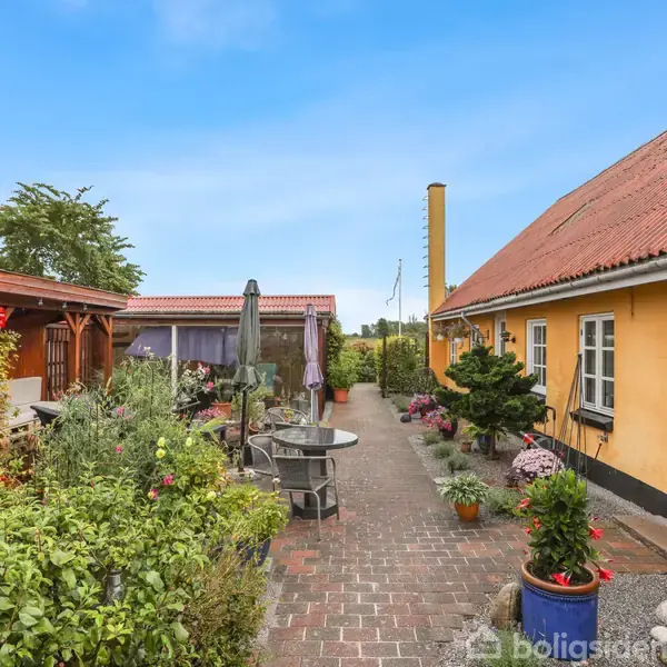 Gårdhave med brostensbelægning omgivet af en gul bygning med rødt tag og en pergola med åbent køkken. Blomster og planter står i krukker langs stien.