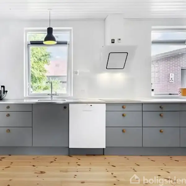 Gråt køkkenbord med indbygget hvid opvaskemaskine og knager, placeret under et vindue med udsigt til grønt område. Over bordet hænger en sort pendellampe.