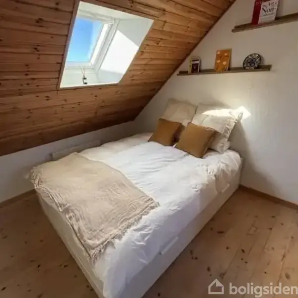 Seng står under skrå loft med træbeklædning, to puder og tæppe på. Ovenlysvindue over sengen. Hylde med bøger og dekorationer på væggen.