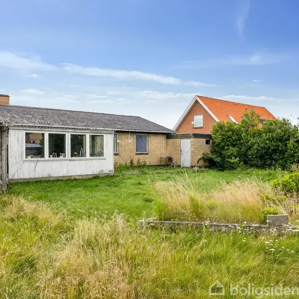 En hvidmalet bygning står stille i en grøn, uplejet have. Ved siden af ligger et hus med rødt tag. Baggrunden indeholder en blå himmel og buske.