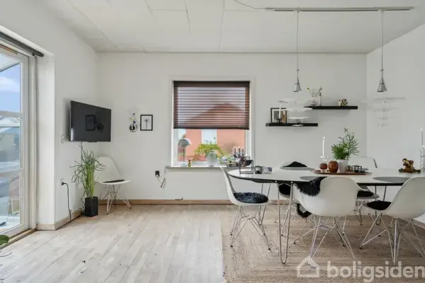 Et moderne spiseområde med rundt bord og seks stole står i et lyst rum. Et vindue med persienner viser en bygning udenfor. En fladskærm er vægmonteret til venstre.