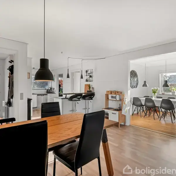 Spisebord med sorte stole står i et åbent køkken-alrum. Et moderne rum med hængende lamper, lyst trægulv og adgang til en spisestue med flere sorte stole og dekoration.
