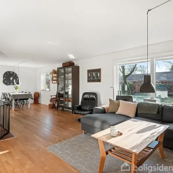 Stue med en sort hjørnesofa står foran et sofabord. En brændeovn er tændt ved en grøn væg. Store vinduer giver udsigt til en have fyldt med træer. En pendellampe hænger over bordet.