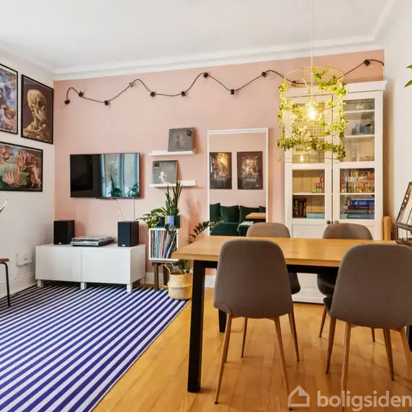 En spisestue med et træbord og fire stole. Væggene har billeder og lysdekoration. En hvid reol med planter og en tv-skærm står ved siden af en blåstribet gulvtæppe.