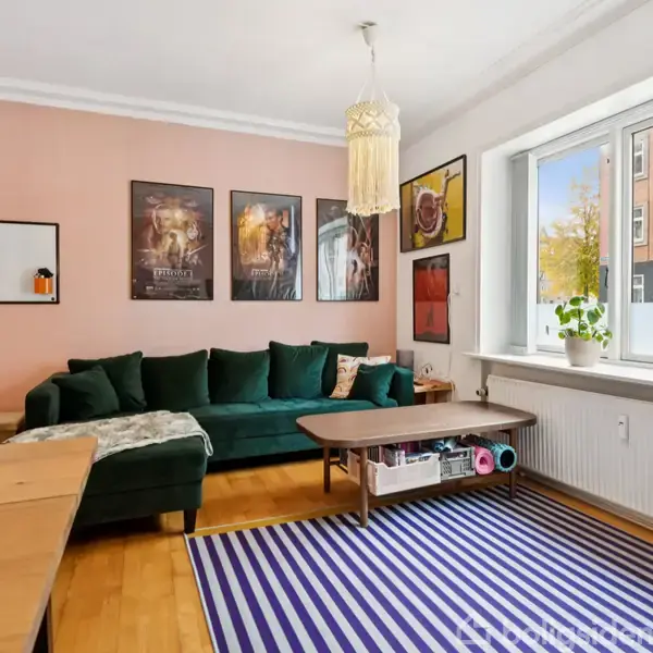 Grøn sofa står i stuen ved siden af et træbord med stribet tæppe og planter i vindueskarmen. Væggene har plakater og en pendellampe hænger fra loftet.