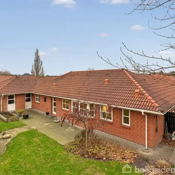 Rødt murstenshus med rødbrunt tag står i en have med grøn græsplæne og flisebelagt terrasse. Træer uden blade omkranser området, og himlen er blå med få skyer.