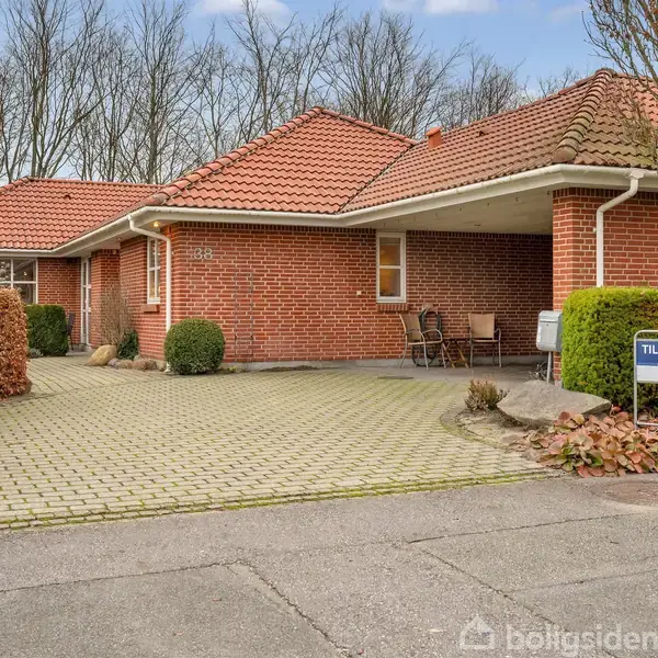 Rød murstenshus med rødt tegltag står stille på en brostensbelagt indkørsel. Til højre ses et "Til salg" skilt. Omkring er der hække og bare træer i baggrunden.