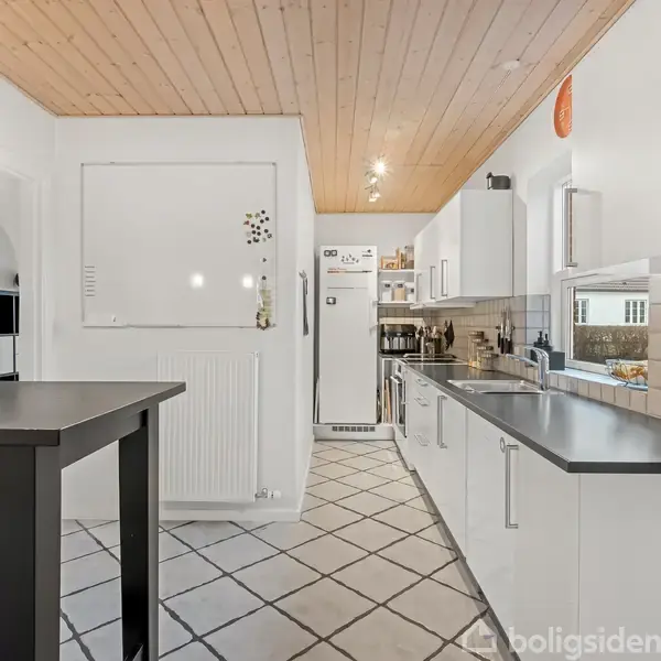 Køkkenbordet står i et lyst køkken med hvide skabe og fliser. En sort spisebord med stole står til venstre. Træloft og lys flisegulv giver en moderne atmosfære.