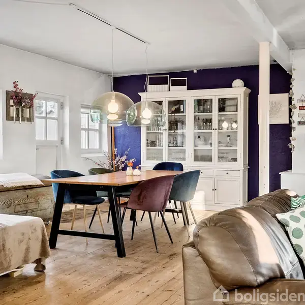 Spisebord med stole står i et lyst rum med et blåt skab i baggrunden. En brændeovn til højre skaber varme ved siden af en grå sofa med pudebetræk med prikker.