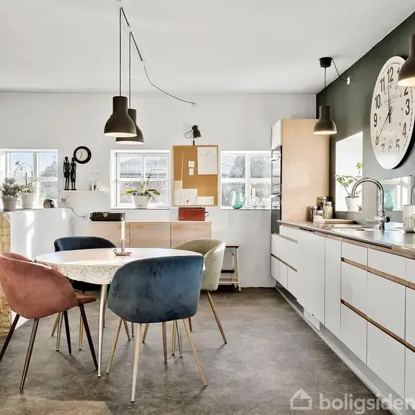 Ovalt spisebord med kulørte stole står under tre lamper i et moderne køkken med hvidt inventar. Stor urskive hænger på væggen. Rummelig stue ses til venstre.