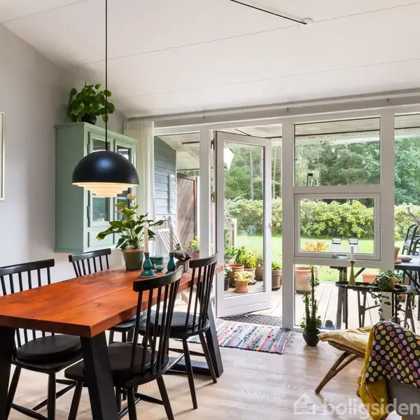 Spisebord med sorte stole står ved glasdøre, der åbner til en terrasse med planter. En vægplakat hænger til venstre. Rummet er lyst med træmøbler og lamper.