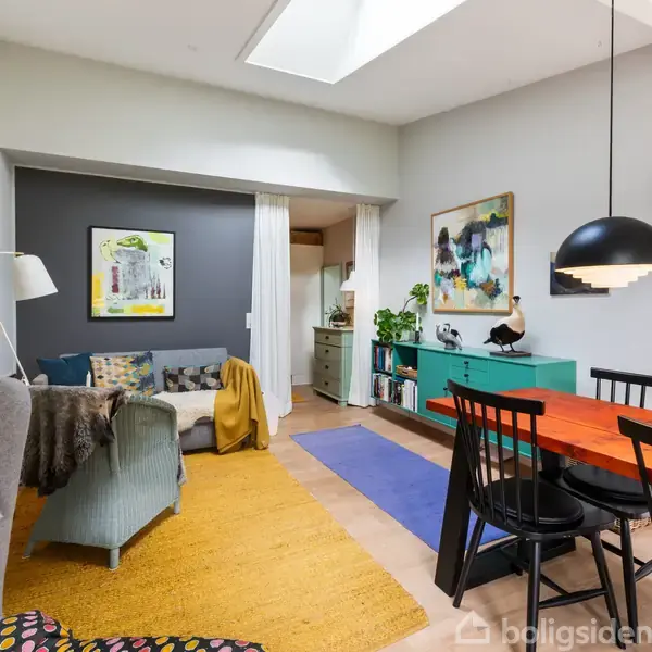 Stue med farverig indretning; sofa og stol til venstre, spisebord med sorte stole til højre. Vægkunst, grøn kommode og planter i baggrunden. Tæpper på gulvet.