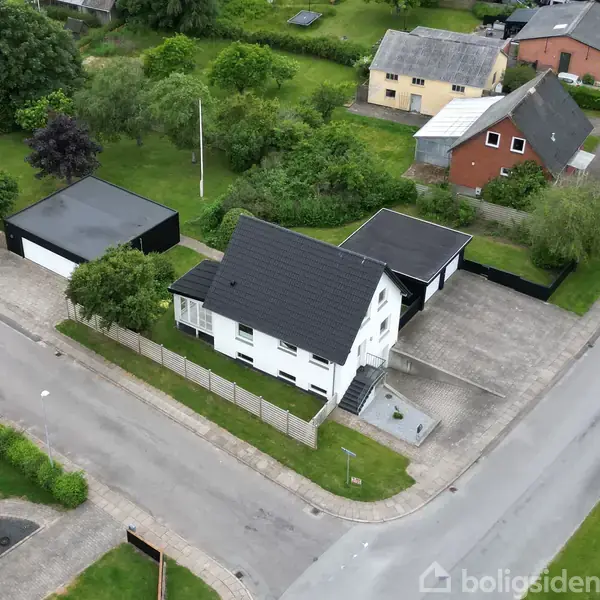 En hvid, to-etagers hus med sort tag står på en hjørnegrund. Ved siden af er en stor garage. Omgivelserne inkluderer andre huse og en grøn have.