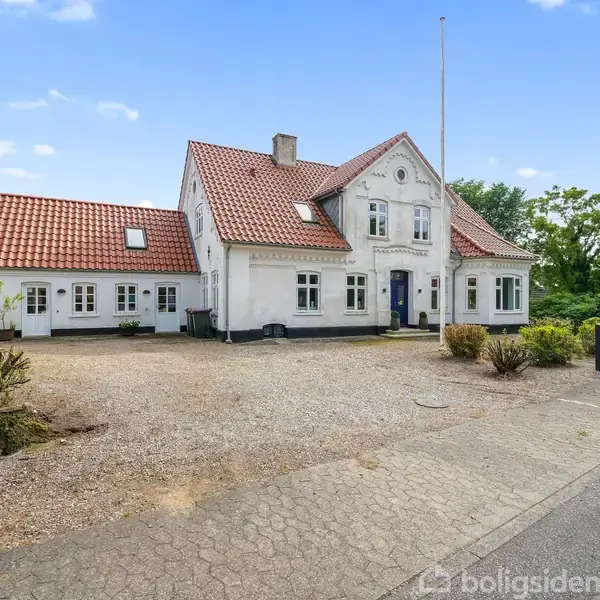 En hvid bygning med rødt tegltag står stille på en grusbelagt grund. Området er belagt med fliser og omgivet af grønne buske og træer, himlen er klar og blå.