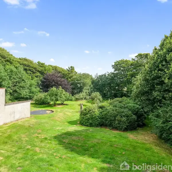 Græsplæne med buske og træer omgivet af en skovbevokset baggrund. En betonmur er til venstre, og himlen er blå med få skyer.
