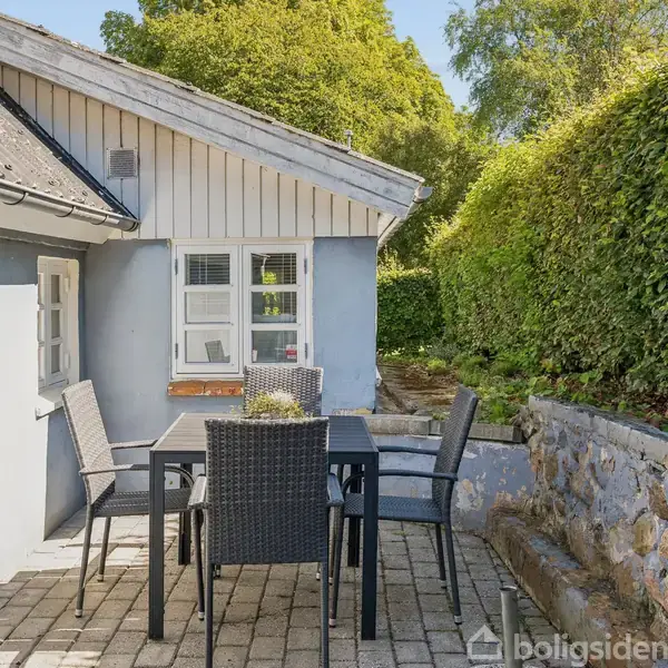 Terrassemøbler står på en stenbelagt terrasse ved siden af et hus med lysegrå vægge. En stensætning og hæk omkranser området. Sollys skinner på møblerne og omgivelserne.