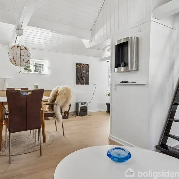 Et spisebord med stole er centralt placeret, dekoreret med fåreskind. Rummet har hvide vægge og bjælker samt en stige til et loftsområde. Møbler inkluderer et vitrineskab.