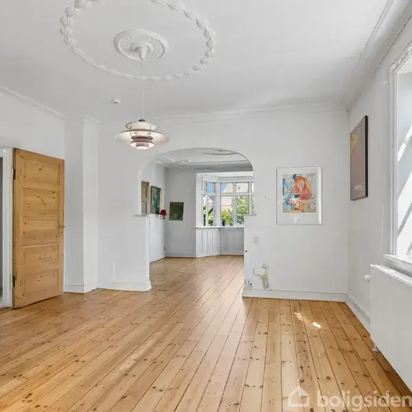 Et lyst rum med trægulv og hvidmalede vægge. Døren står åben til venstre, en rund loftsroset med lampe ses centralt, og vinduer slipper lys ind fra højre.