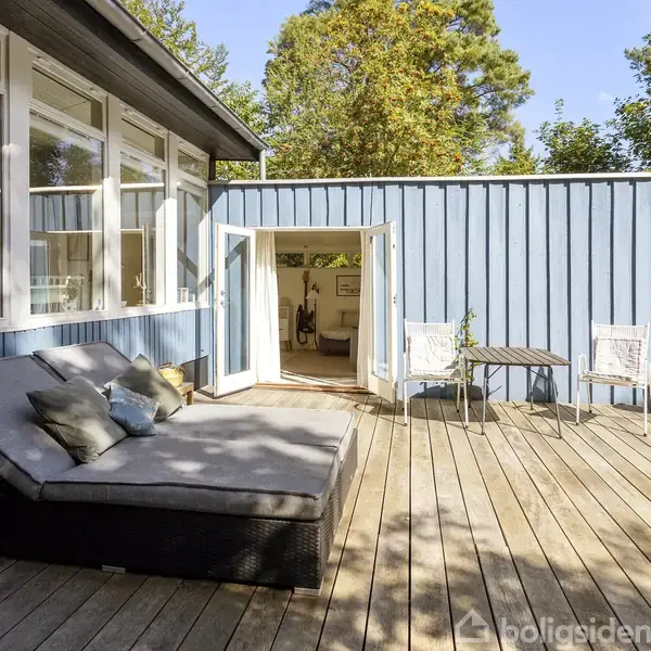 En solrig træterrasse har en stor, komfortabel daybed med puder. To hvide stole står ved et lille bord nær åbne dobbeltdøre, der leder ind til et lyst rum.