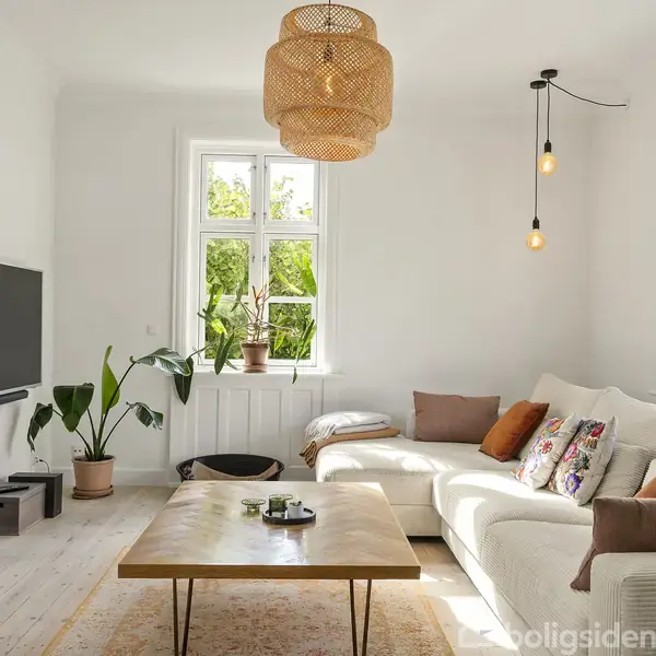Tv hænger på væggen, mens en stor, hvid sofa med farverige puder står ved vinduet. Træbord i midten. Grønne planter dekorerer det lyse rum, oplyst af en stor loftslampe.