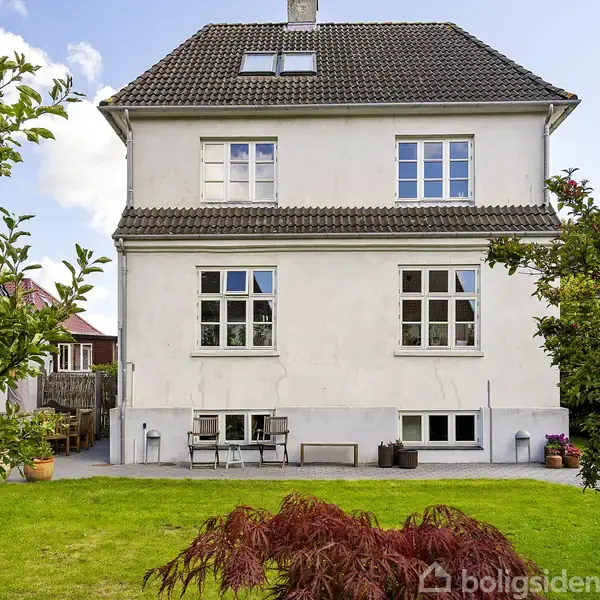Et hvidt hus med brun tagsten står i haven, omkranset af grøn græsplæne og buske. To store vinduer ses på første sal.
