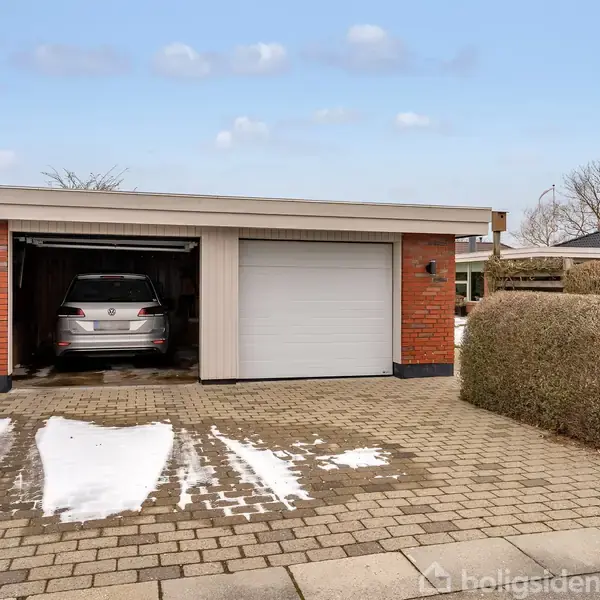 Garage bygning med en bil inde, åben port til venstre, lukket port til højre. Mursten og træstruktur. Omgivet af snedækkede fliser og klippede hække.
