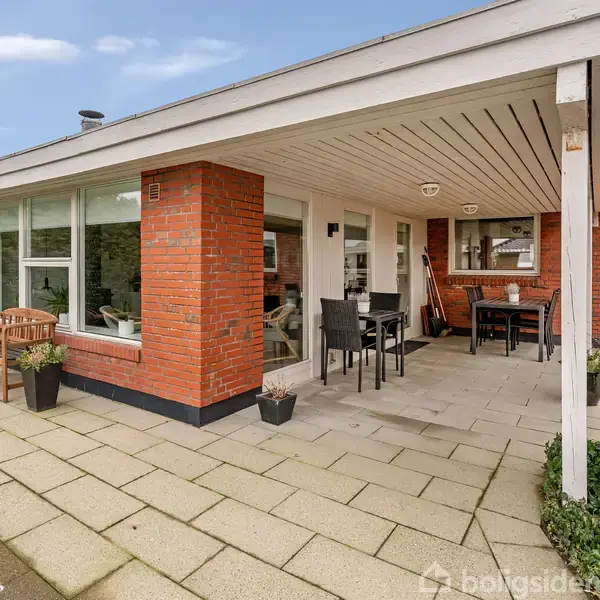 Rødstenshus med overdækket terrasse har udendørs borde og stole. Omgivet af flisebelagt område og grønne planter.